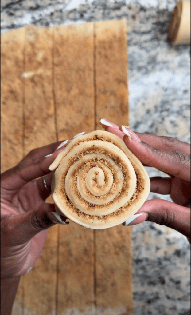 Close-up of hands holding a cinnamon roll dough with a spiral shape and a sprinkle of cinnamon sugar on top, set against a granite countertop.