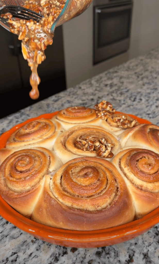 A bowl pouring a caramel mixture over freshly baked cinnamon rolls topped with nuts, placed in a round orange dish on a granite countertop.