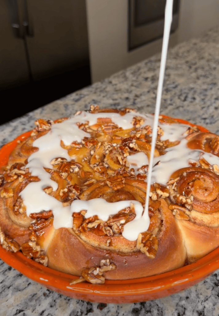 A cinnamon roll dessert topped with cream cheese frosting and chopped pecans, placed in a deep orange dish on a granite countertop.