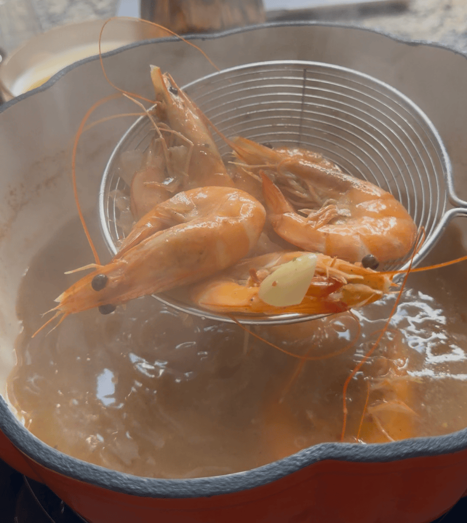 A strainer lifting cooked shrimp from a pot of steaming broth, revealing their vibrant orange color and a hint of garlic.