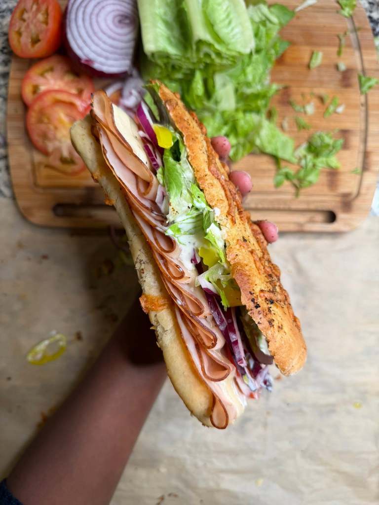 A hand holding a freshly made sandwich loaded with layers of deli meats, lettuce, and red onions, with sliced tomatoes and a head of lettuce in the background on a wooden cutting board.