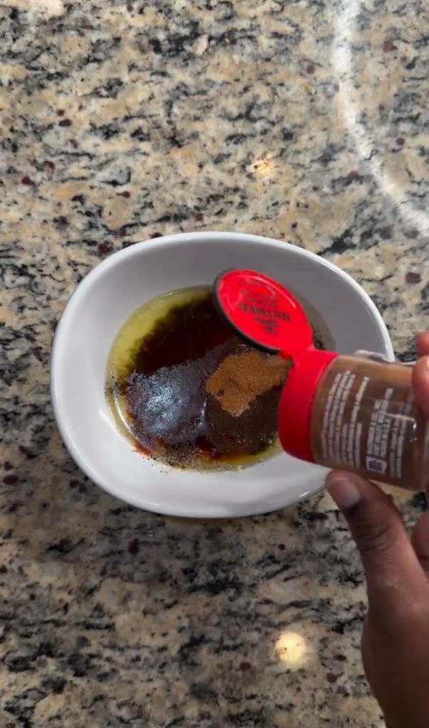 A hand pouring a red spice from a container into a bowl of dressing on a granite countertop.