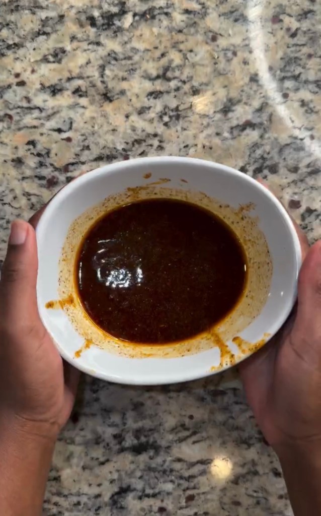 A person's hands holding a white bowl filled with a dark, thick sauce on a granite countertop.