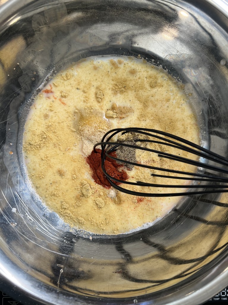 A stainless steel bowl containing a mixture of eggs, spices, and milk, with a whisk stirring the ingredients.