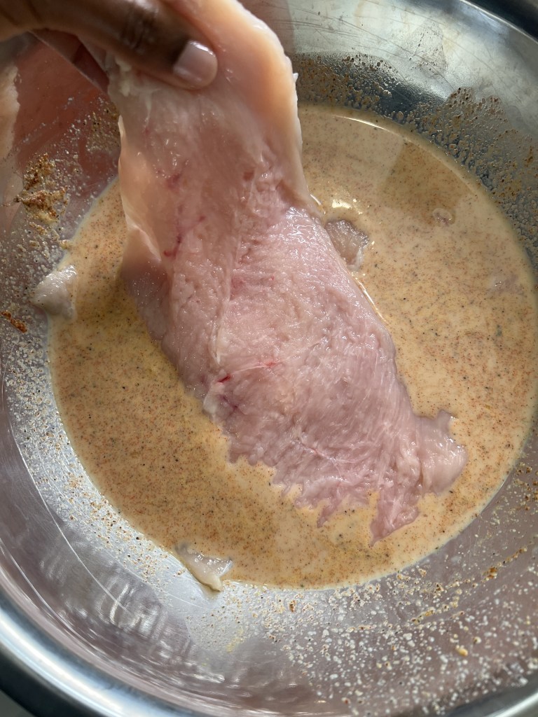 A close-up image of a hand holding a piece of raw chicken breast submerged in a creamy marinade inside a stainless steel bowl.