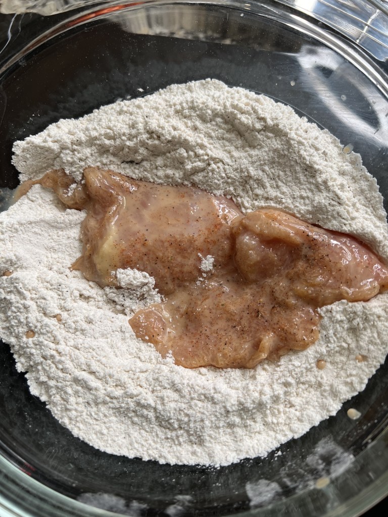 Raw chicken pieces coated in seasoned flour in a glass bowl.