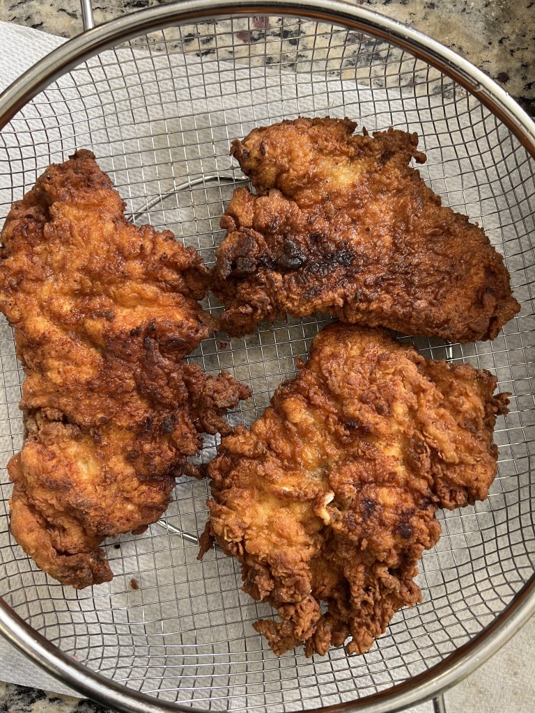 Three pieces of crispy fried chicken resting in a wire mesh strainer on a countertop.