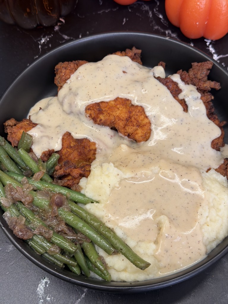 A plate of Southern fried chicken covered in creamy gravy, served with mashed potatoes and green beans.