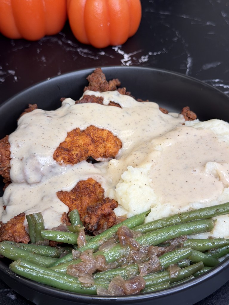 A plate of fried chicken topped with creamy gravy, served with mashed potatoes and green beans. Two orange pumpkins are in the background.